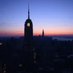 Widok panoramy miasta o zmierzchu z najwyższym budynkiem, Empire State Building, w centrum. Wschodzące słońce maluje niebo w odcieniach różu i pomarańczu, tworząc piękny kontrast z granatowym niebem nad głową.